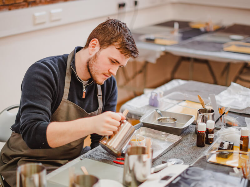 A person sits at a candle making class, pouring molten wax into a container